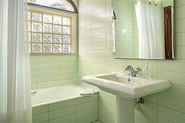 Pale green tiled bathroom featuring a bathtub, white shower curtain, pedestal sink with faucet, and a glass block window with an arched top.