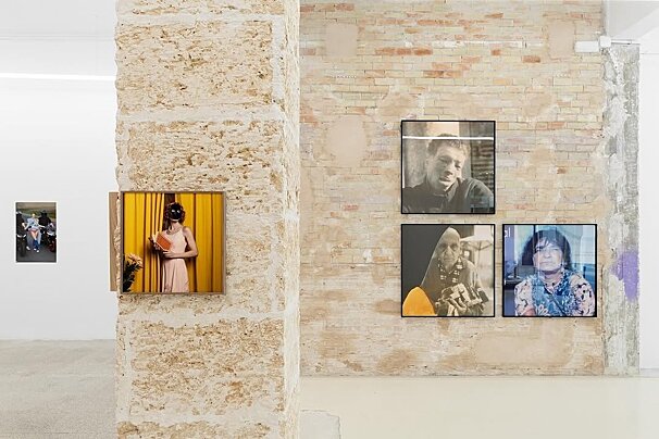 An art gallery displays various framed photographs on textured stone and brick walls, featuring portraits and a masked figure holding a book.