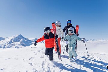 A happy family of four in ski gear walks playfully on a snowy mountain peak under a clear blue sky.