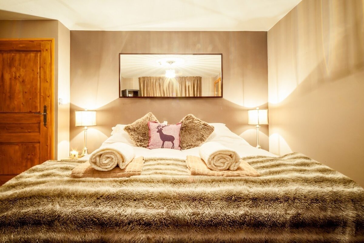 A warm and inviting bedroom features a large bed with a faux fur blanket, a deer-themed pillow, bedside lamps, and a mirror.
