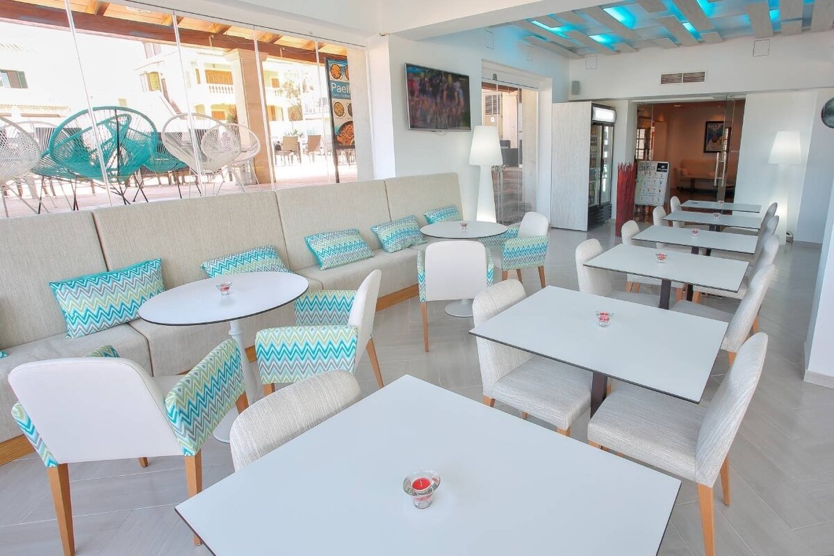 Bright, modern cafe interior with white tables, beige bench seating with turquoise chevron cushions, and a glass wall overlooking an outdoor patio.