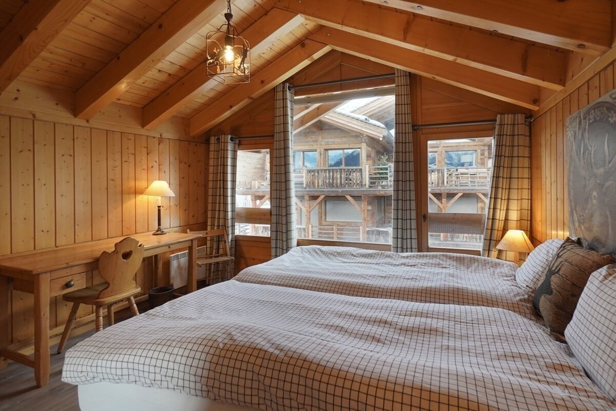 Cozy, wood-paneled bedroom with two beds, a desk, and a rustic chalet view through attic windows.