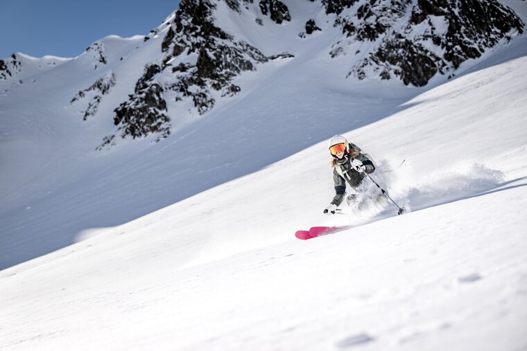 A skier on pink skis carves a turn down a vast, snowy mountain, kicking up powder. Sunlit peaks loom under a blue sky.