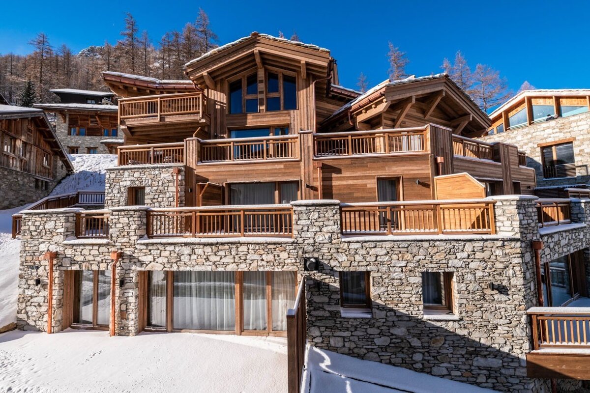 A luxurious, multi-level wooden and stone ski chalet with balconies, nestled in a snowy mountain landscape under a bright blue sky.