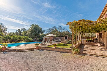 An inviting villa patio with a long swimming pool, sun loungers, stone architecture, and a leafy pergola under a bright blue sky.
