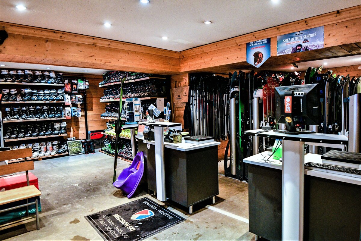 A busy ski and snowboard rental shop with wooden walls, featuring shelves of boots, racks of skis/boards, and several service counters.