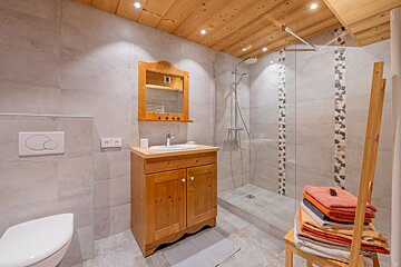 A bright bathroom features light gray tiles, a wooden ceiling, a wood vanity, mirror, toilet, and a glass shower with mosaic details. Towels are stacked.