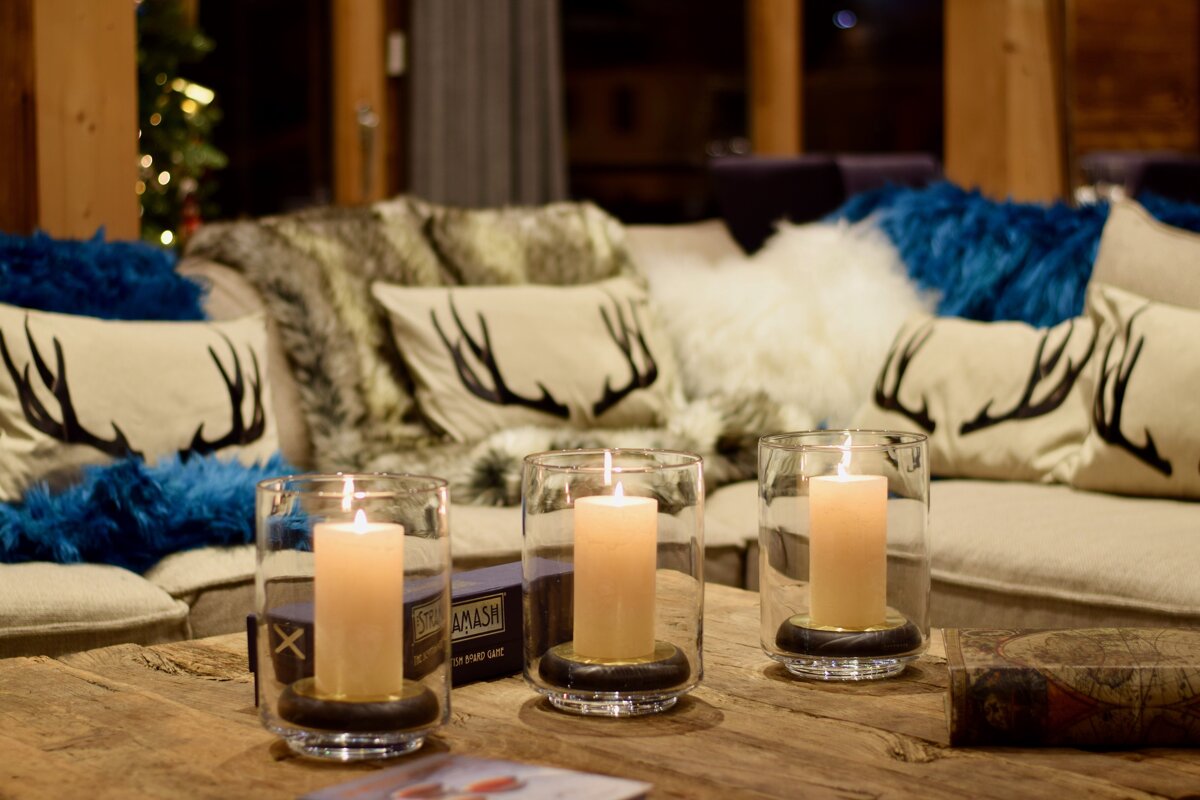 Three lit candles on a wooden table, with cozy couches adorned with antler pillows and fluffy throws in a warm, inviting living room setting.