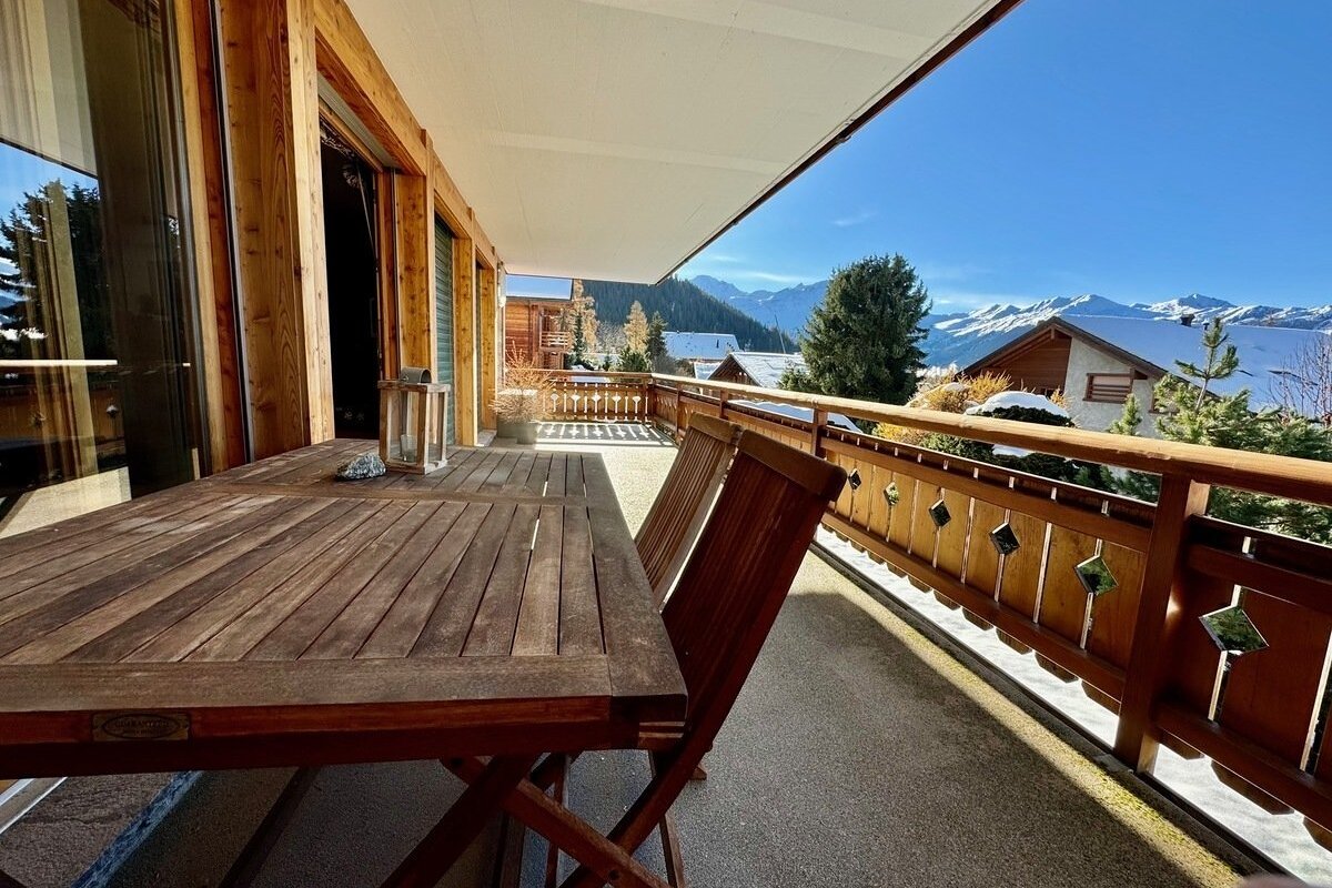 A sunlit balcony with a wooden table and chairs, offering a stunning view of snowy mountains and a winter village under a clear blue sky.