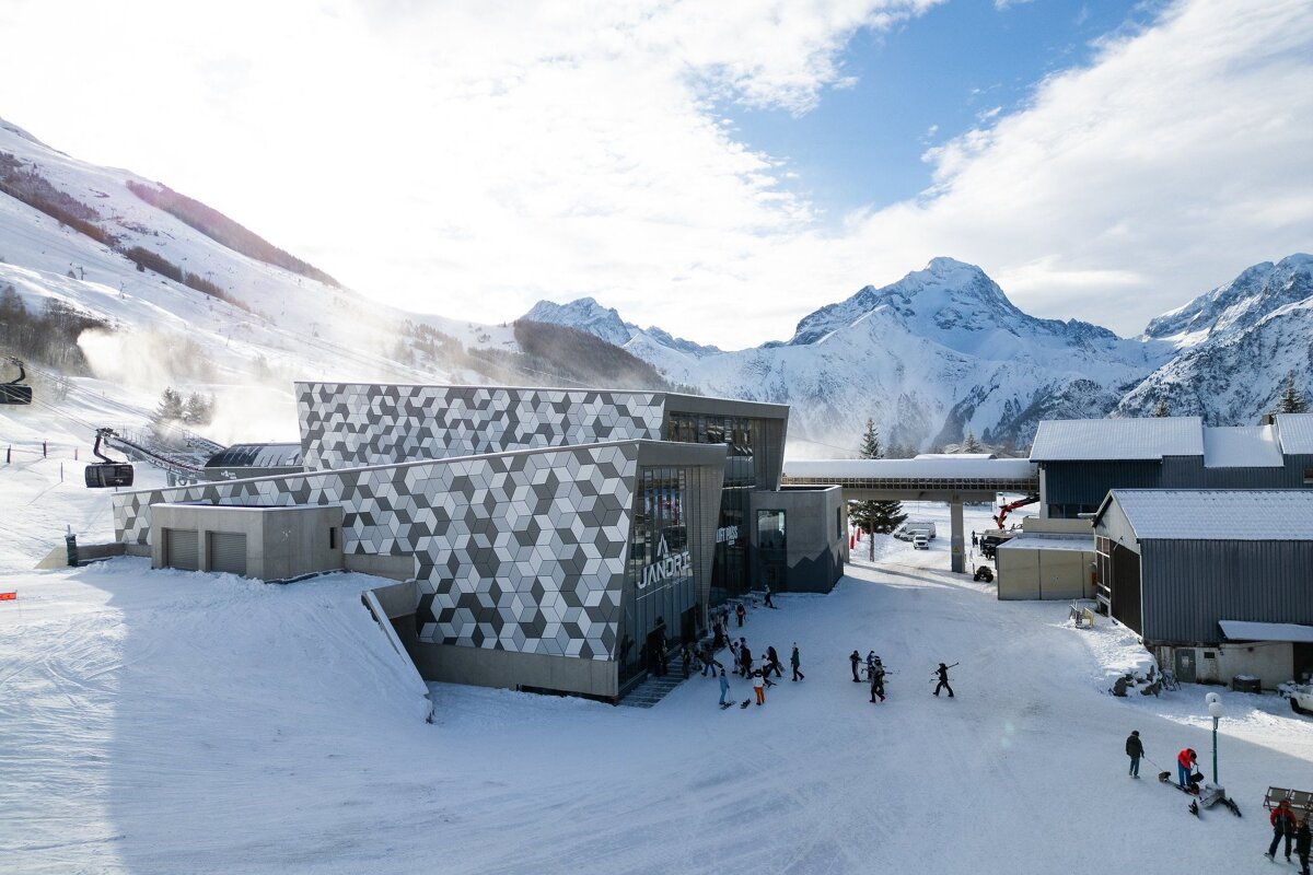 A stylish ski resort building with a geometric pattern sits in a snowy mountain landscape. Skiers and a lift are visible under a bright sky.
