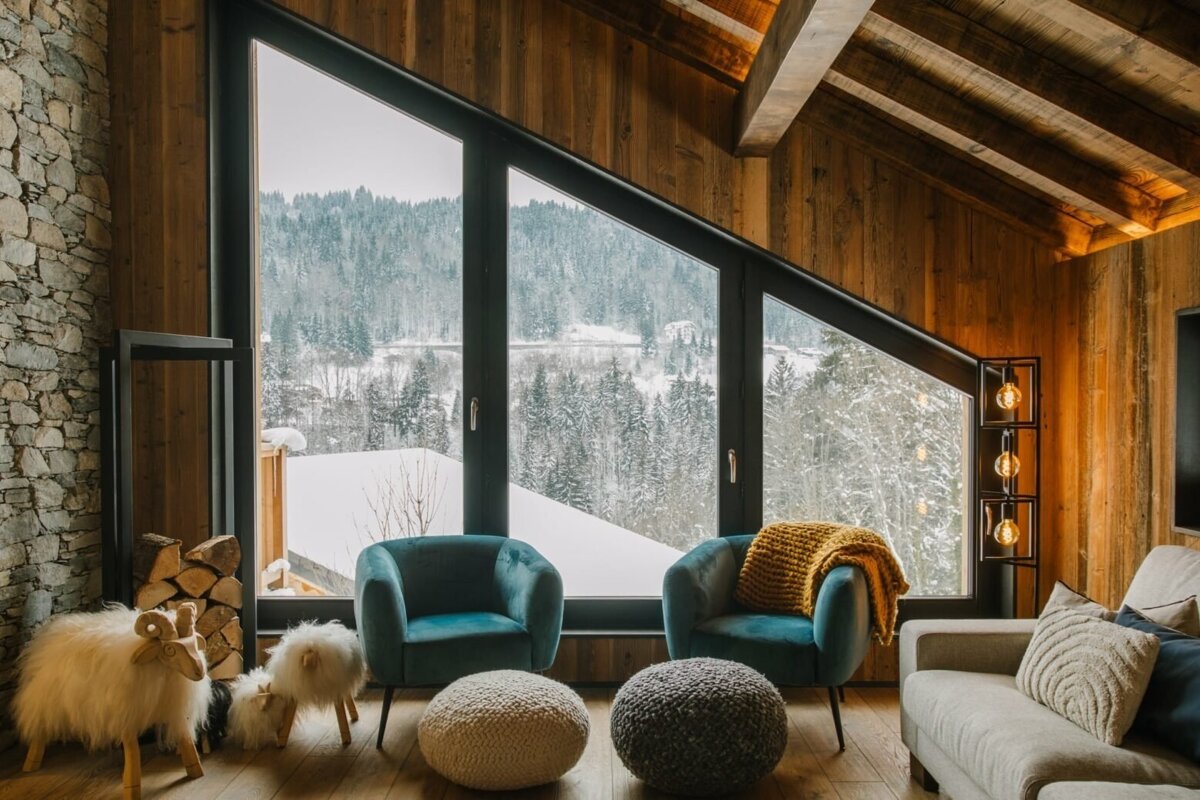 A cozy, modern alpine living room features large angled windows showcasing a snowy forest view. Rustic wood and stone accents complement plush seating and warm lighting.