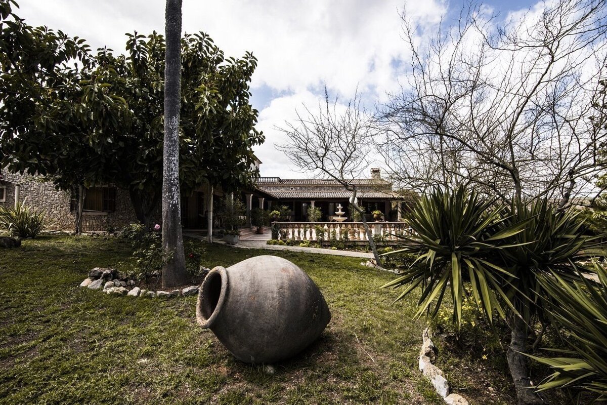 A rustic garden with a large clay pot on grass, leading to a stone house with a tiled roof, surrounded by diverse foliage under a cloudy sky.