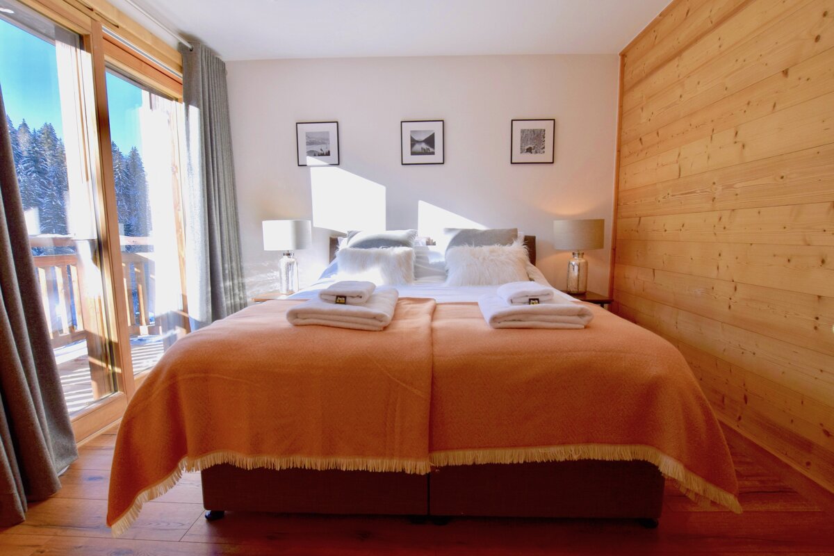 Bright, cozy bedroom with an orange blanket, large bed, and a balcony view of snowy mountains. Features wooden floors and paneling for a warm, ski-lodge feel.
