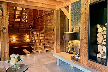 A cozy chalet interior with wood-paneled walls, a modern staircase, stone accent wall, and a niche filled with firewood.
