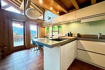 Modern kitchen with white cabinets and island in a rustic wooden chalet. Large windows overlook stunning mountain views. Exposed beams & tiled floor.