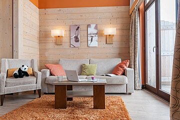 Cozy alpine-style living room featuring a beige sofa, a panda plush on an armchair, a coffee table with a laptop, and an orange rug.