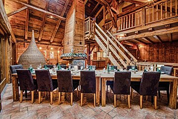A warm, rustic wooden dining hall features a long table set for dinner, a large fireplace, exposed beams, and a mezzanine, creating a cozy lodge atmosphere.