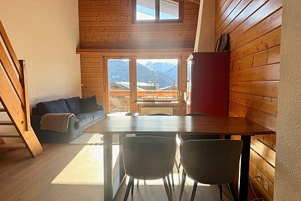 A sunlit chalet living room with wood paneling, a dining table, sofa, and large windows offering a beautiful snowy mountain view.