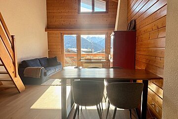 A sunlit chalet living room with wood paneling, a dining table, sofa, and large windows offering a beautiful snowy mountain view.