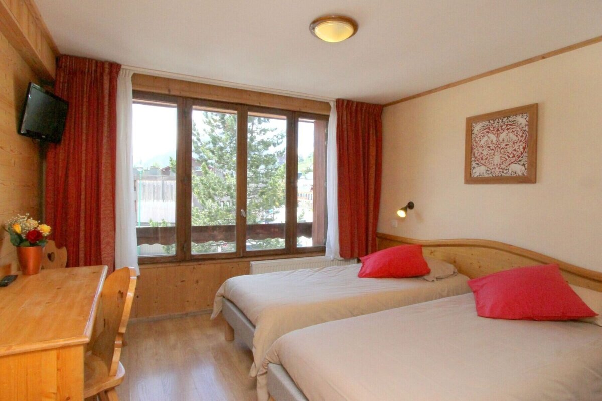 A cozy bedroom with two single beds, red pillows, a wooden desk, and a wall-mounted TV. A large window overlooks trees, complemented by red curtains.