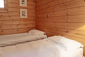 Two single beds with white bedding in a room featuring light wood-paneled walls, framed art, and a small window.