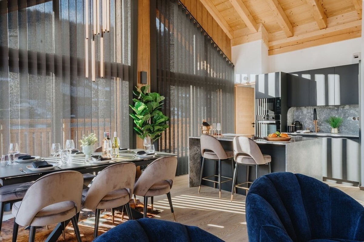 A chic, sunlit modern interior with an open-plan dining area and kitchen, high wood-beamed ceilings, and large windows casting stripes of light.