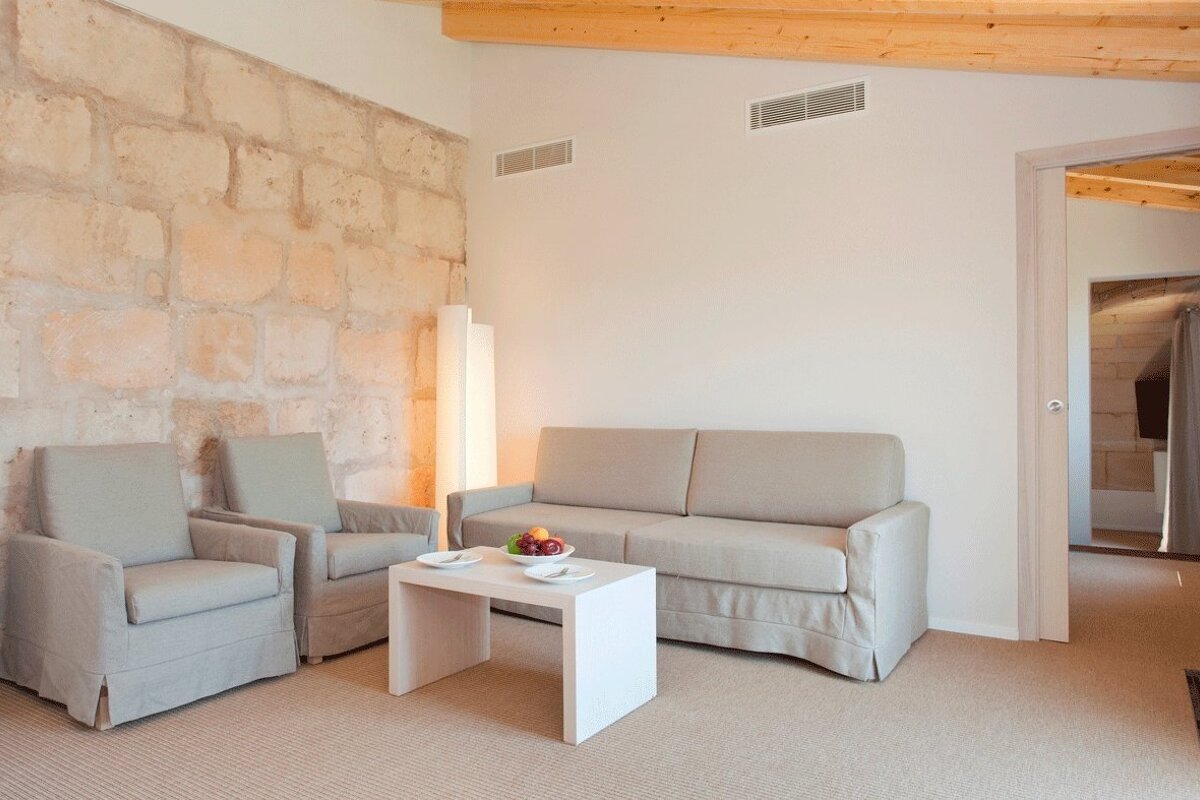 A modern living room featuring a stone accent wall, beige sofa, two matching armchairs, and a white coffee table with a fruit bowl. Wooden beams are visible.