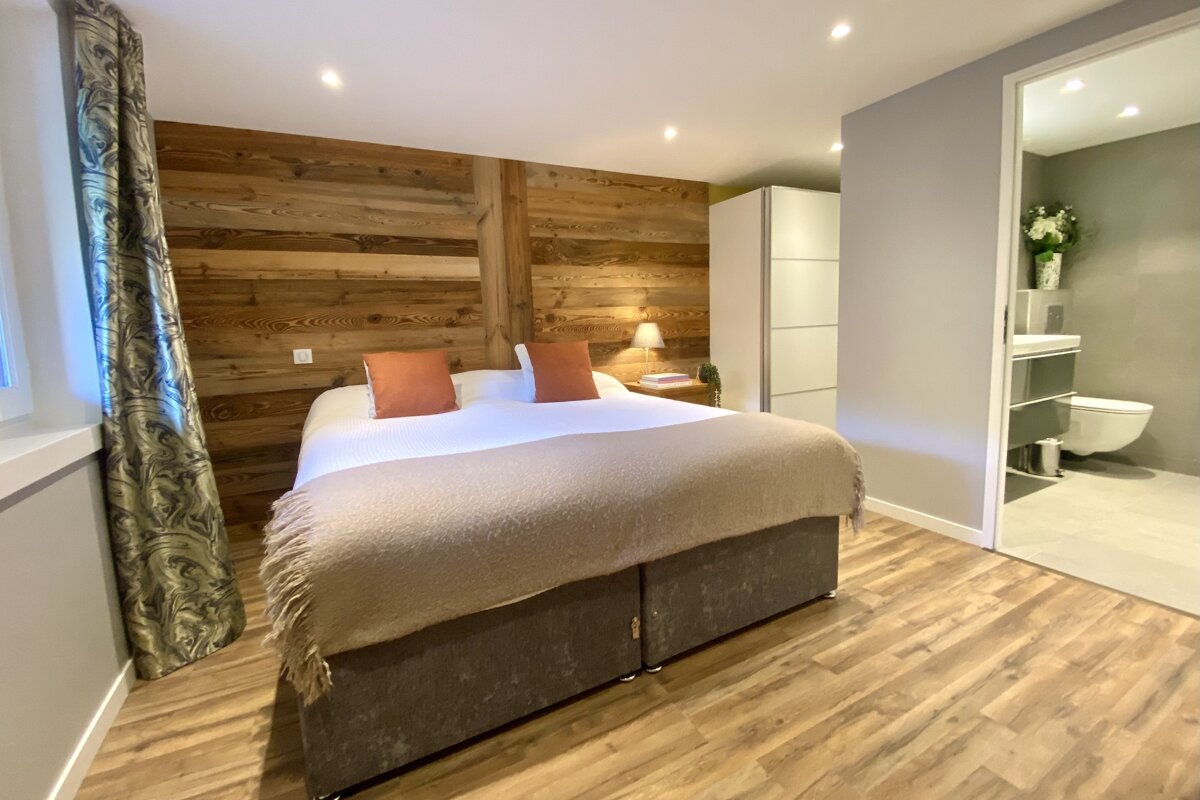 A chic bedroom features a large bed, rustic wood paneling, and a modern ensuite bathroom visible through an open doorway.
