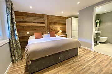 A chic bedroom features a large bed, rustic wood paneling, and a modern ensuite bathroom visible through an open doorway.