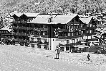 Black and white image of a snowy ski resort. Hotel Champs Fleuris overlooks a slope with two skiers. Snow-covered mountains and chalets fill the background.