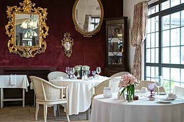 An elegant dining room with rich burgundy walls, ornate gold mirrors, white-clothed tables, wicker chairs, and a large window.