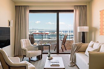An elegant living room opens to a balcony with panoramic views of a sunny marina, filled with boats and a red cargo ship.