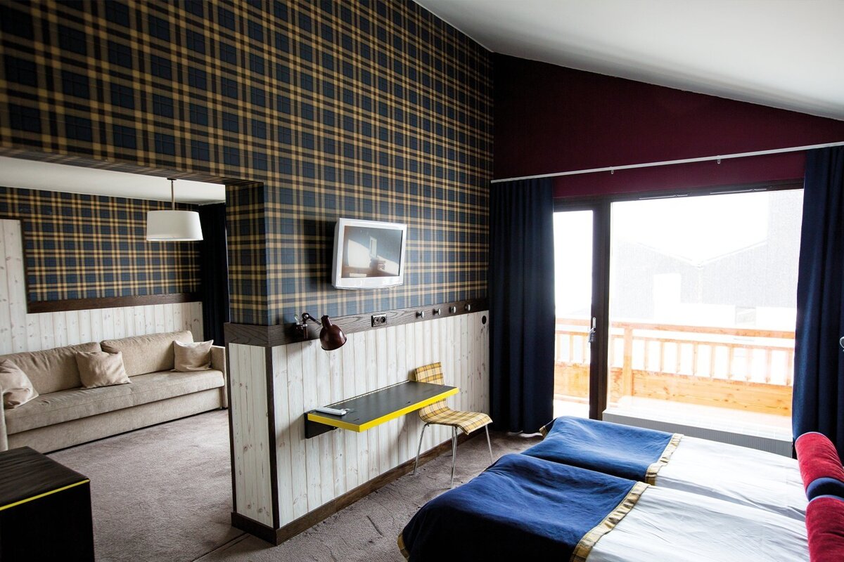 A cozy hotel room with dark blue and gold plaid wallpaper, wooden paneling, a seating area with a sofa, twin beds, and a balcony door.