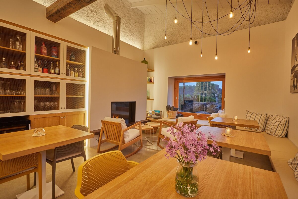A cozy, modern dining/lounge area with a fireplace, unique hanging lights, wooden tables, a bar, and a large window overlooking greenery.