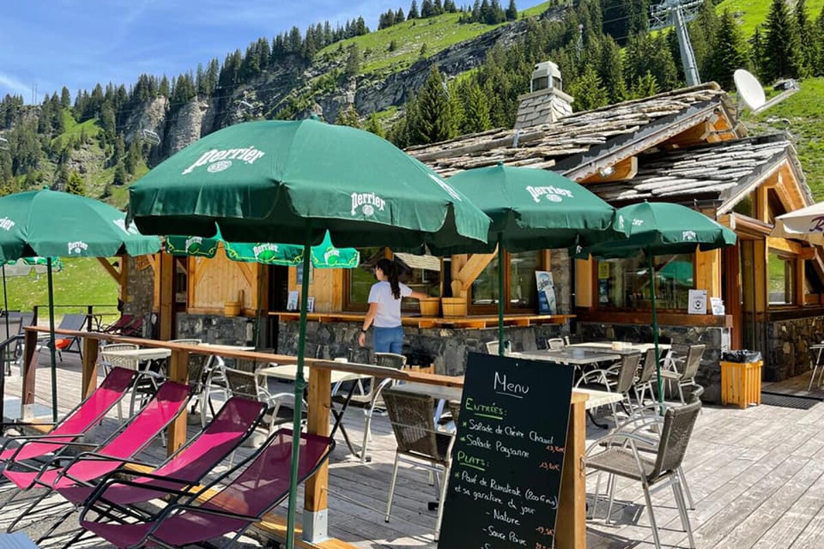 A sunny mountain restaurant terrace with green Perrier umbrellas, outdoor seating, and lounge chairs, set against a backdrop of green mountains.