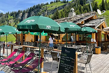 A sunny mountain restaurant terrace with green Perrier umbrellas, outdoor seating, and lounge chairs, set against a backdrop of green mountains.