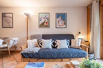 A cozy living room features a blue futon with patterned pillows, Chamonix ski posters, a warm floor lamp, and wooden furniture.