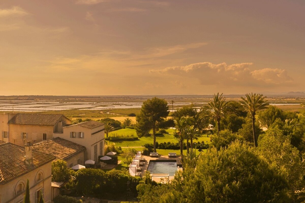 Elevated view of a villa with a pool, surrounded by lush gardens and palm trees, overlooking distant salt pans and flatlands under a golden sky.