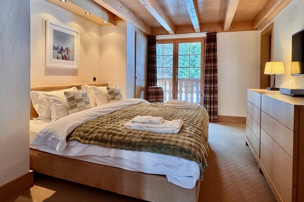 Cozy, rustic bedroom with a large bed, wooden ceiling beams, balcony access, and a dresser. Warm lighting highlights the comfortable ambiance.