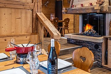 A cozy, rustic chalet dining scene with a fondue pot, wine bottle, and a warm, glowing fireplace.