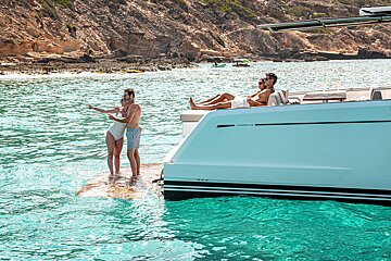 People relaxing on a luxury yacht in turquoise water near a rocky shore, with some sunbathing and others on the swim platform.