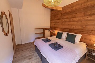 A cozy bedroom with a large bed, prominent wooden accent wall, and light wood flooring. A woven ceiling light and wooden mirror add character.