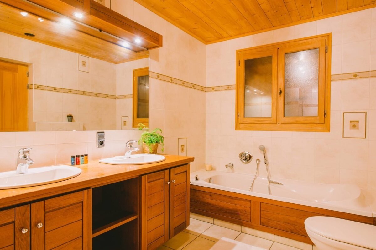 A warm, inviting bathroom featuring double sinks on a wooden vanity, a large mirror, a built-in bathtub, and warm wood ceilings and window frames.
