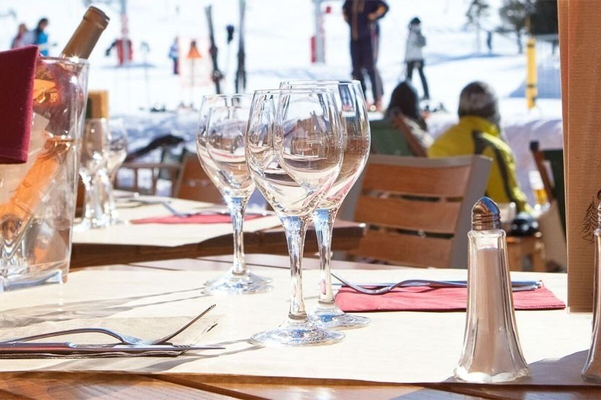 A table set with wine glasses and a wine bottle in an ice bucket outdoors at a sunny ski resort restaurant, with skiers in the snowy background.