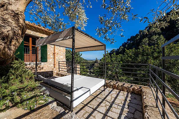 A cozy outdoor canopy bed on a stone patio overlooks a lush green valley and mountains under a bright blue sky, beside a rustic stone house.