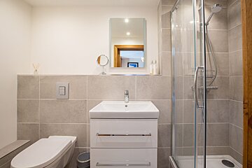 A modern bathroom features a white toilet, vanity with sink, and glass shower enclosure. Neutral grey tiles and white walls create a clean aesthetic.