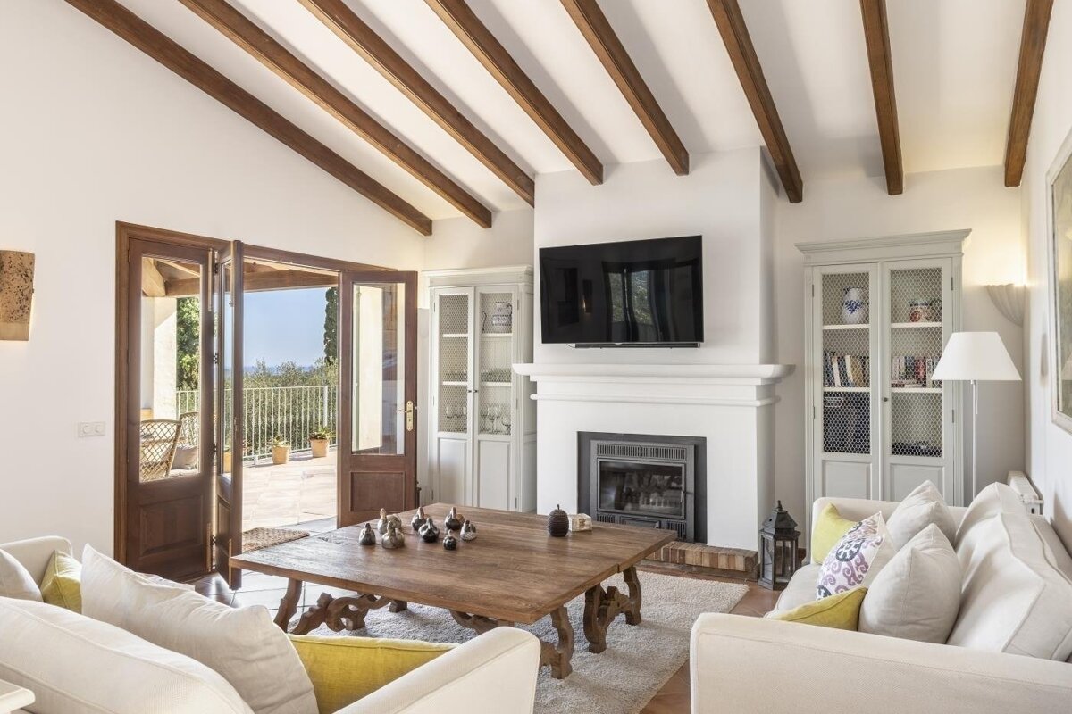 A bright, rustic-chic living room with vaulted beamed ceilings, white sofas, a large wooden table, and French doors opening to a scenic outdoor terrace.