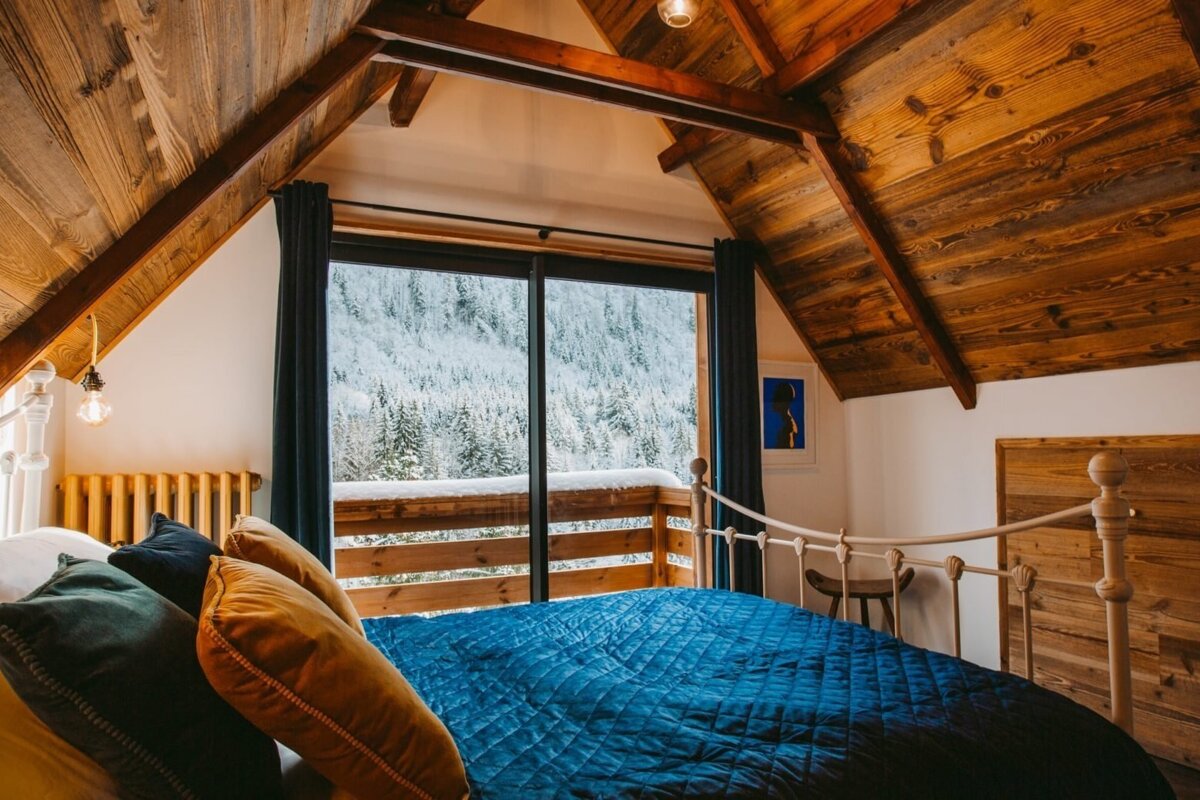 Cozy attic bedroom with a blue bedspread, wooden ceilings, and a large window offering a stunning view of snow-covered mountains and pine trees.