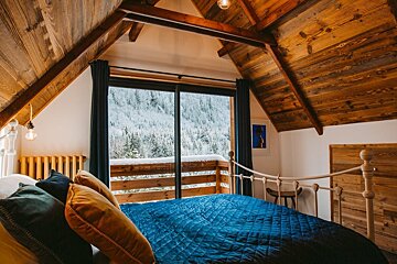 Cozy attic bedroom with a blue bedspread, wooden ceilings, and a large window offering a stunning view of snow-covered mountains and pine trees.