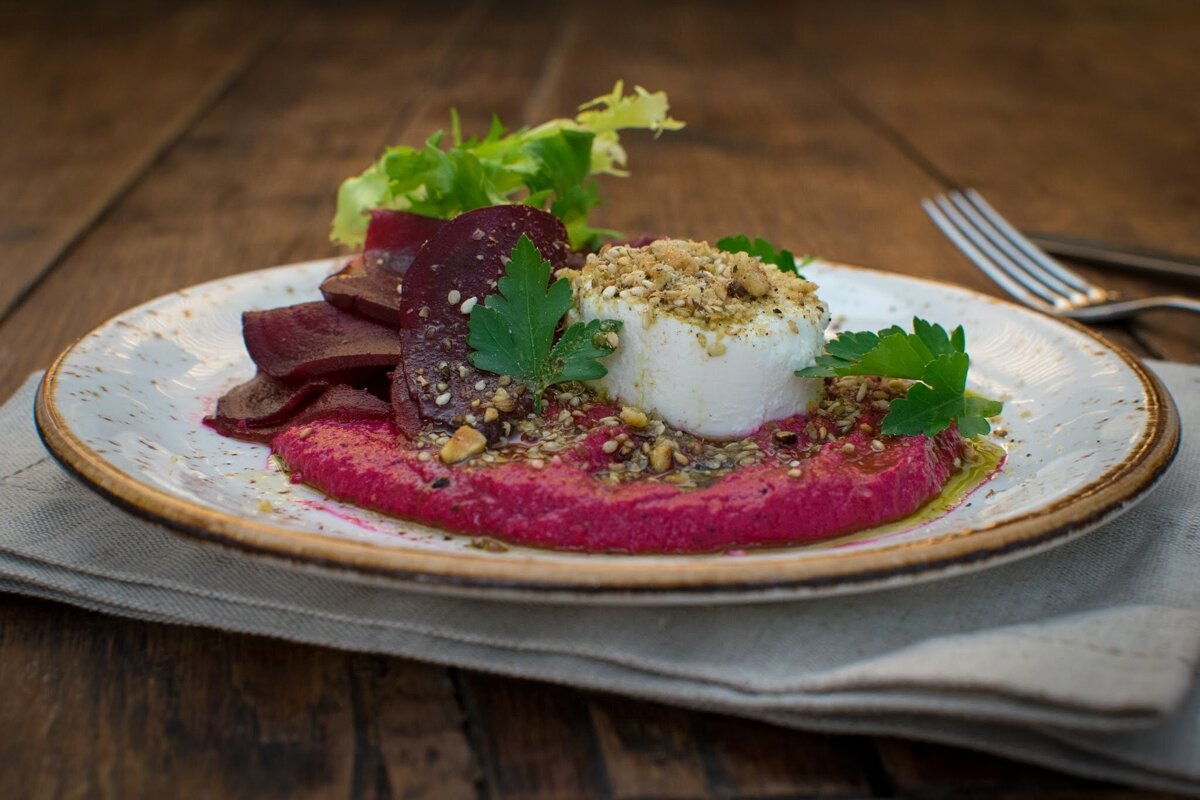 An elegant dish of goat cheese atop bright beetroot hummus with sliced beets, herbs, and a nut sprinkle on a rustic plate.